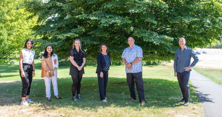 Royal Roads University president stands with former and current students in front of a big Linden tree.