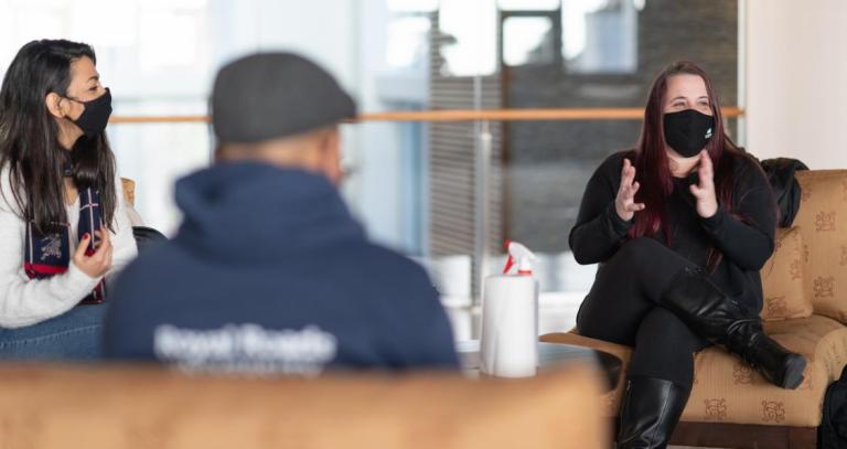 Three students sit in an open lobby. They are physically distanced and wearing masks, but quite engaged in what one students is saying.