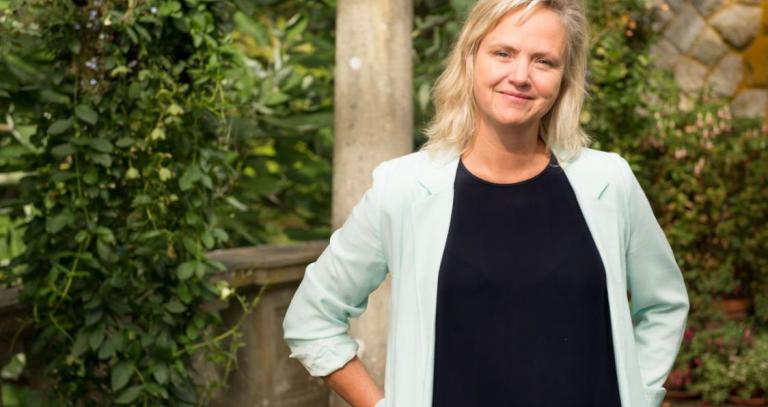 A smiling woman with blonde, shoulder-length hair wearing a mint green blazer and black t-shirt standing in front of a background of green leaves and stone columns.