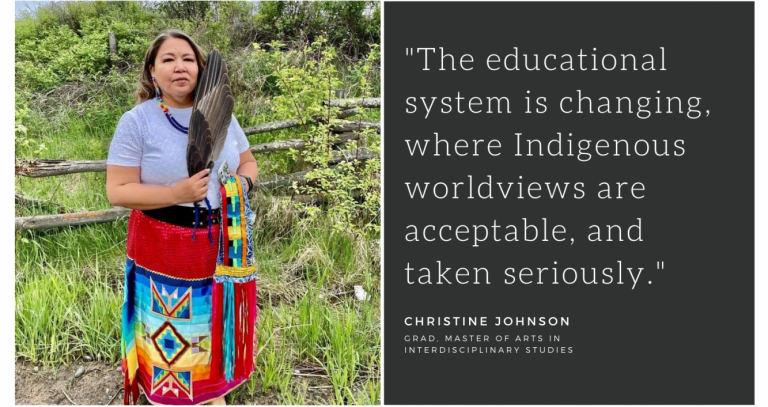 Christine Johnson stands in front of green trees and bushes. She is holding a full wing and is dressed in a bright red skirt and a beaded necklace.