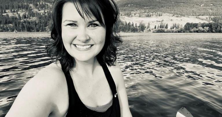 Black and white photo of a smiling woman with mid-length dark hair taken on a kayak from the middle of a lake. A treed and hilly landscape is in the background.