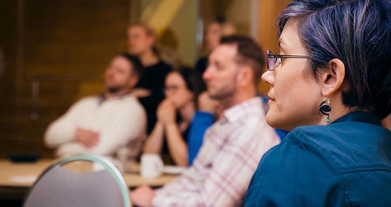 Prof. Jaigris Hodson studiously watches a presenter at a conference. Her purple hair glints in her glasses. 