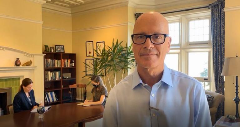 President Steenkamp in the foreground of his bright office. Behind him, two staff members work at a big table and chat. One is wearing a mask.