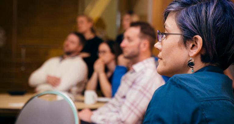 A group of people seated in a room, their attention is focused toward an (unseen) presenter. A woman with dark hair streaked with purple is in the foreground.