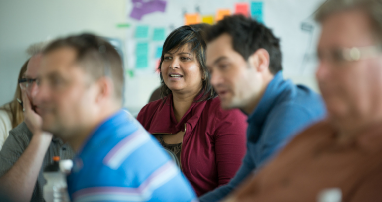 A group of MBA students attend a class at Royal Roads University, a white board with colourful post-it notes in the background.