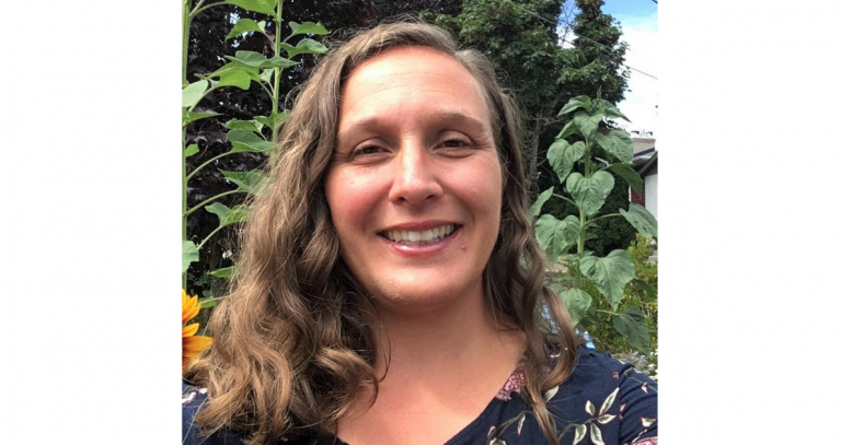 smiling person in front of tall sunflower greens that are above their head and even taller green fir trees in background