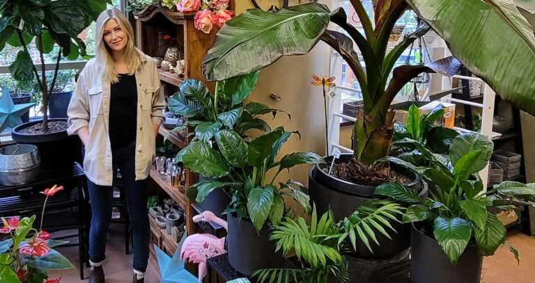 A woman stands in doors surrounded by towering potted plants and leafy greens.