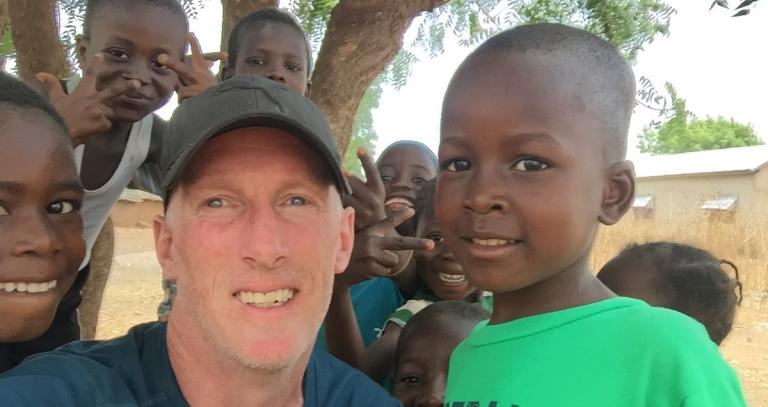 Man in baseball hat squats down so a group of seven children can also be seen, they are gathered around and smiling into the camera