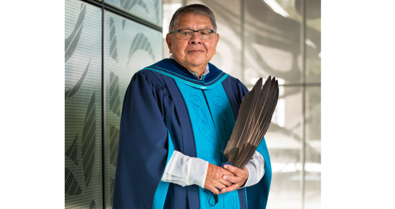 Lekwungen Elder Butch Dick standing in convocation regalia with traditional embroidery down the front, holding eagle feathers. 