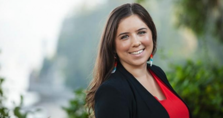 A smiling woman poses for the camera with a blurred out background of bushes and trees behind her.