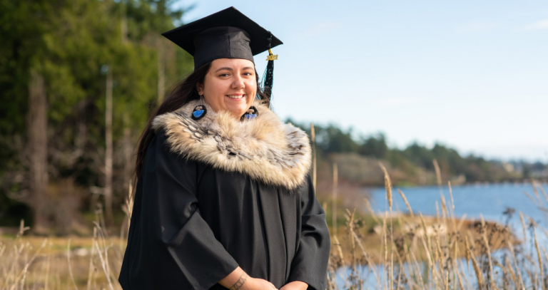 A woman stands in front of a lagoon wearing a graduation cap and gown.