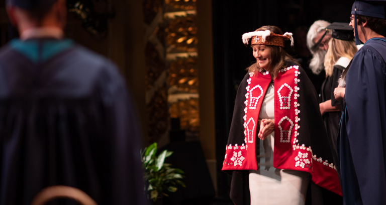 A graduate crosses the convocation stage draped in a red and black button blanket.