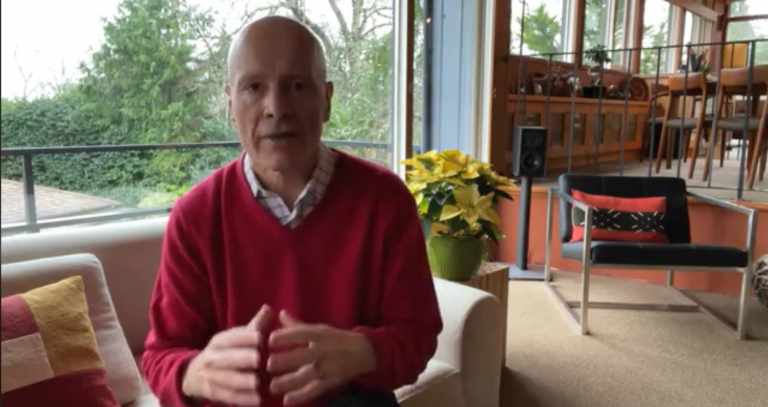 A man sits in his livingroom with a window and potted plant behind him.
