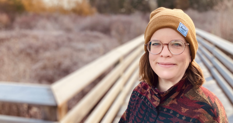 A woman in a toque smiles at the camera while standing on a boardwalk.