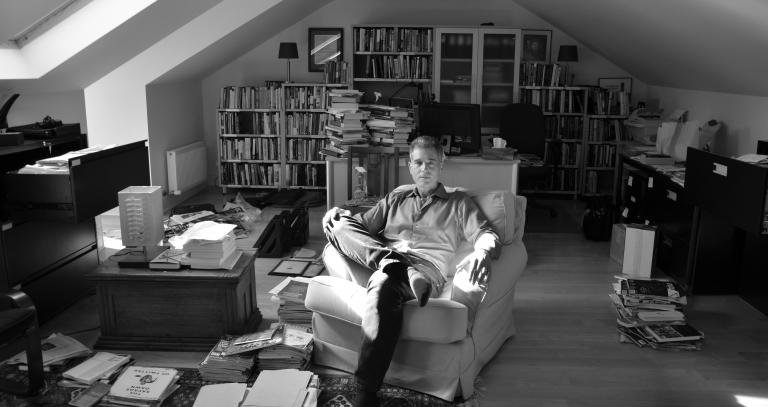 Black and white image. A man sits in a wingback chair surrounded by books. The sun streams in through the window.