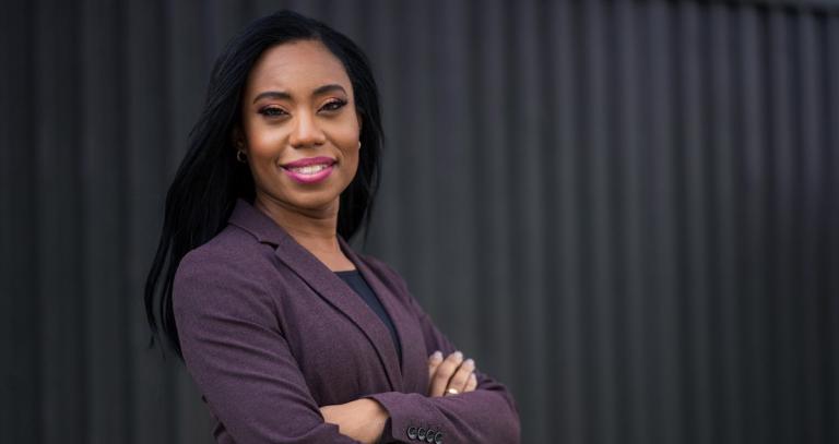 RRU grad Siobhan Calderbank, a Black woman in a purple suit jacket, crosses her arms and smiles. 