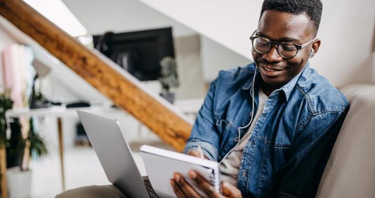 A man smiles while holding a not pad and a laptop computer.