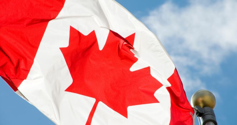 The Canadian flag waves in front of a blue sky with fluffy white clouds.