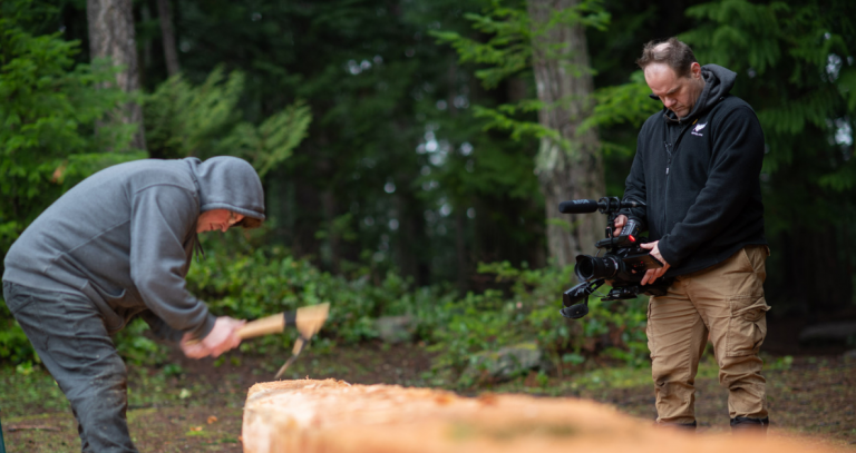 A man holds a video camera to record an Indigenous carver at work. A forested backgroudnd can be seen.