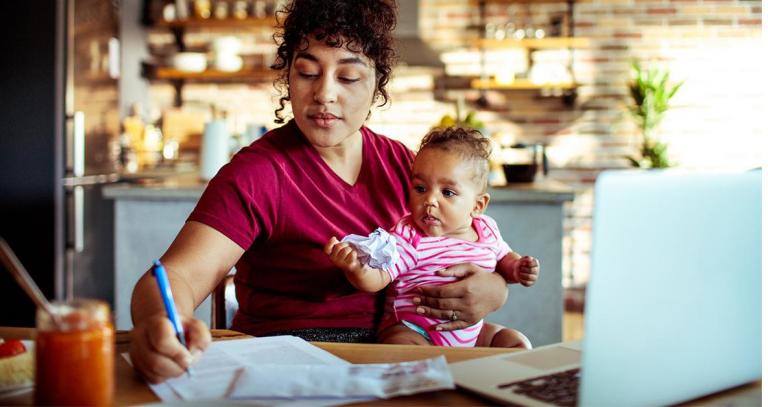 A woman holding a baby while seated at a desk with a laptop and while she writes.