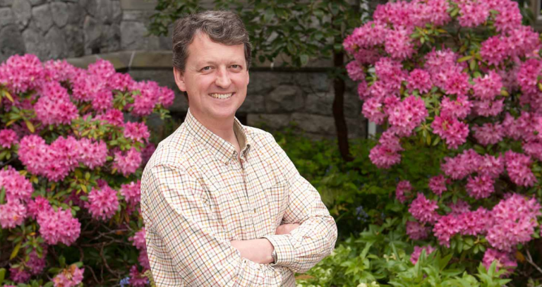 Head shot of Geoff Bird standing in front of a stone wall with bushes filled with purple flowers in front