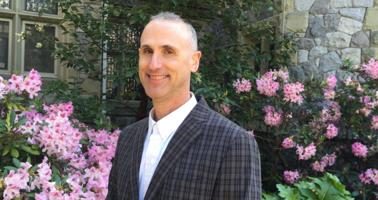 Prof. Geo Tkach smiles in front of flowering shrubs at Hatley Castle