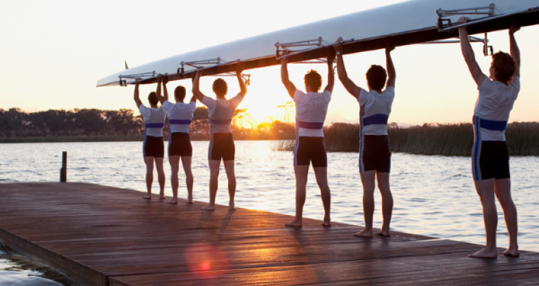 Rowers holding up a boat on a dock with the sun rising in the background