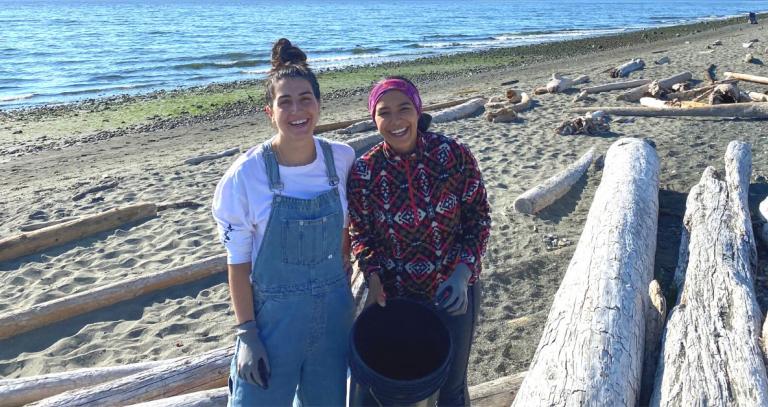 Two BBA students, Emily (left) and Celicia (right) stand on the beach at Esquimalt Lagoon with a bucket for trash