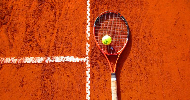 Tennis racket laying on bright orange ground with yellow ball balanced on top of it.