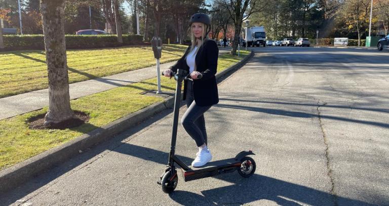 Karly Nygaard-Petersen stands with one foot on an electric scooter on a tree-lined street