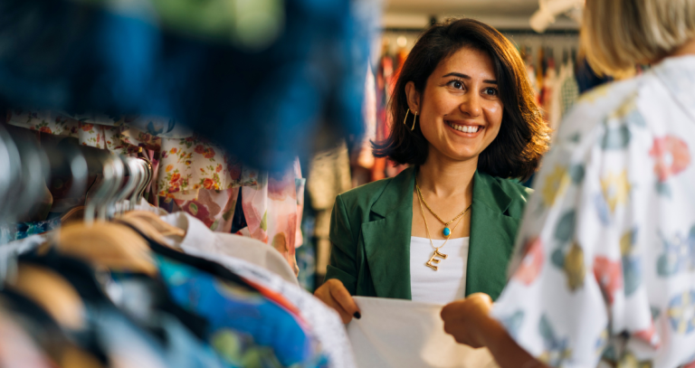 Two women smile at each other while shopping among clothes in a thrift store.