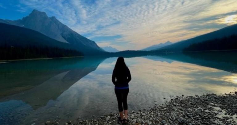 Kate Dunn stands on a rocky beach with a blue lake and mountains behind her