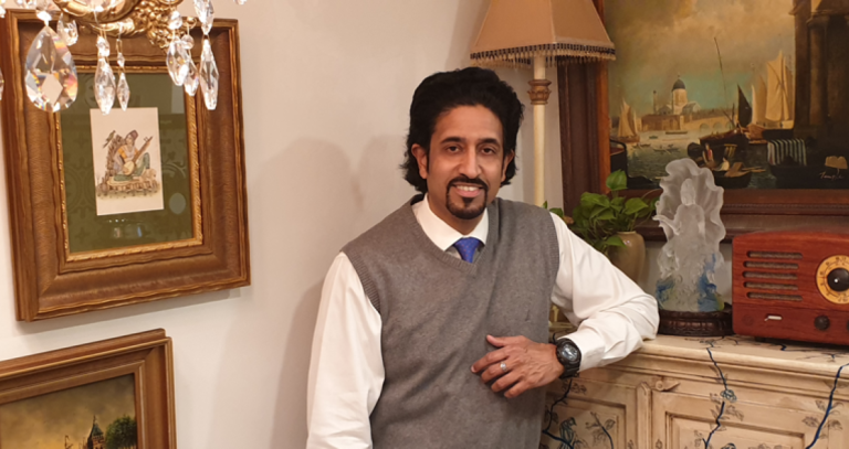 Man wearing sweater vest and dress shirt leans on a piece of furniture. Framed artworks are on the walls.