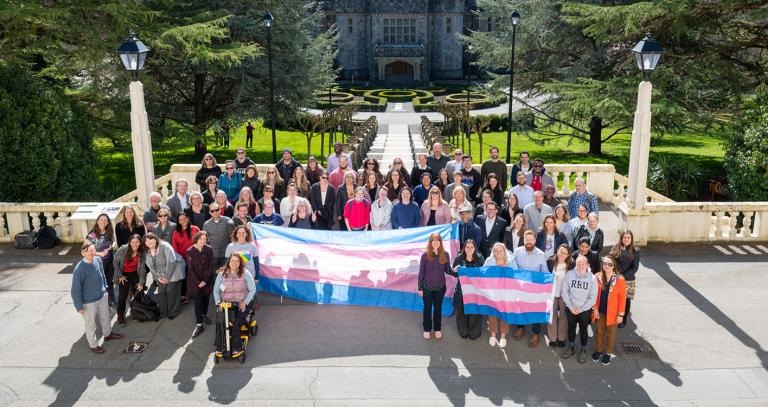 Royal Roads University staff and students pose in front of a long staircase holding up the Transgender flag.