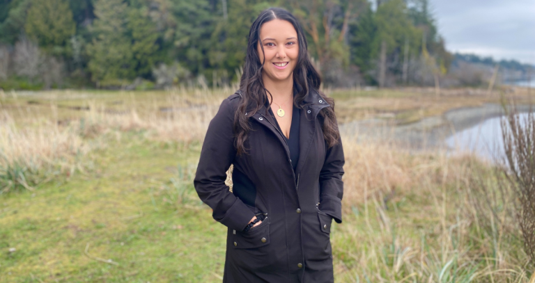 Candice Cook stands in front of an ocean and forested background.