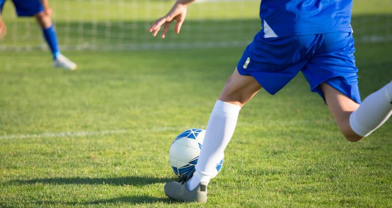 A child in a blue soccer uniform kicking a soccer ball towards a net