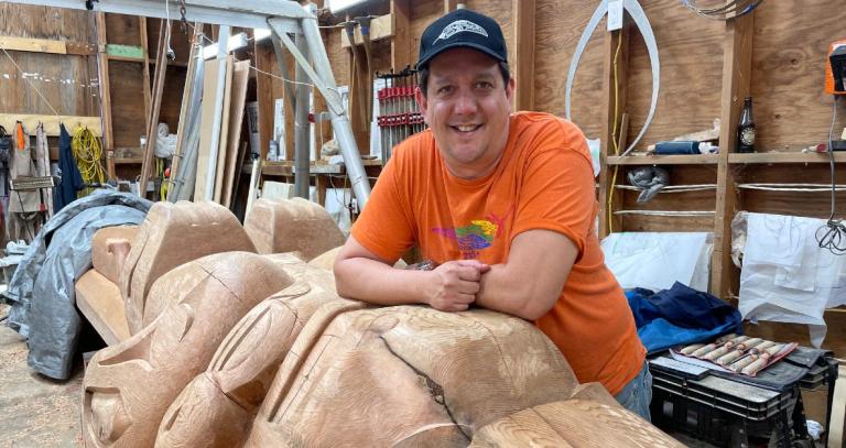 Smiling Indigenous man resting his arms on a partially carved totem pole in a workshop. He is wearing an orange t-shirt and a dark ball cap. 