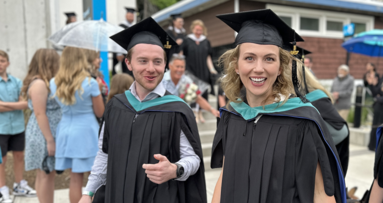 Two graduates whoosh by the camera in their regalia smiling brightly.