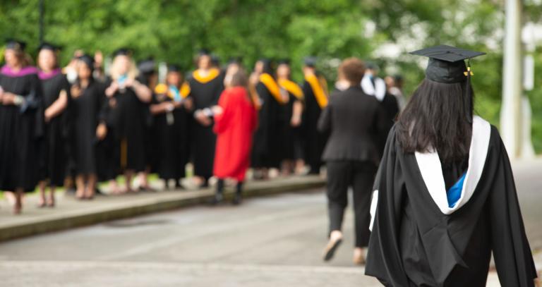 line of grads in robes in the background, with a woman in grad robes in the foreground walking towards them