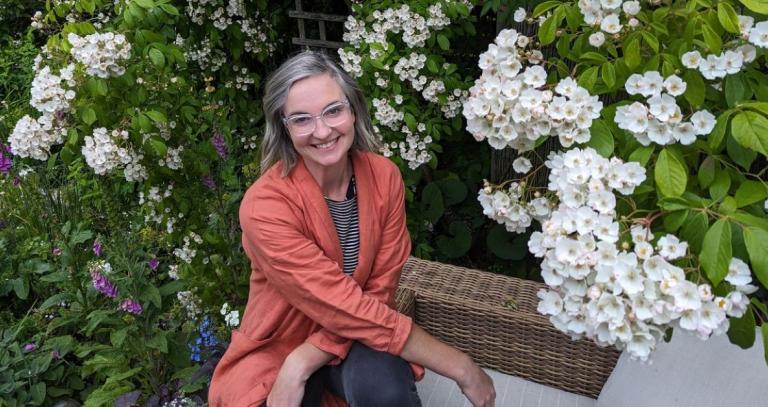 A woman wearing a coral blazer smiles out directly at the camera. She is surrounded by a blooming wisteria 
