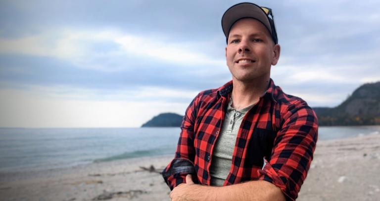 Gary Hayes stands on a beach wearing a flannel shirt and smiling. 