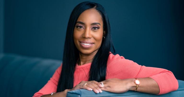 Siobahn Calderbank is a Black woman with long straight black hair. She is seated and leaning over the arm of a velvet blue sofa.