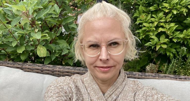 A woman with light blonde hair smiles into the camera in front of a wall of greenery
