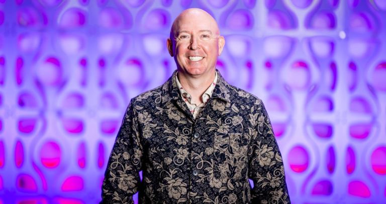 A smiling man wearing a gold patterned onyx jacket smiles out directly at the camera. He is in front of a blurred but vivid purple abstract background.