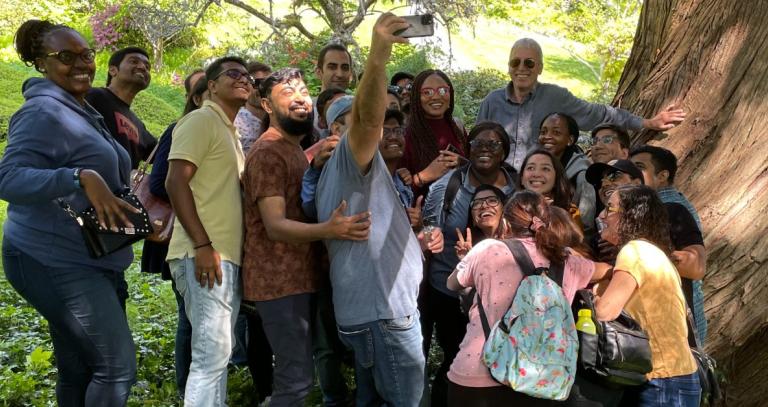 Large group of students surrounding their teacher, taking a selfie by a large tree trunk.