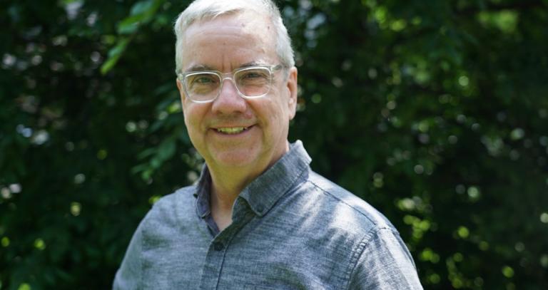 Man with grey hair, glasses and wearing a blue shirt smiling at the camera.