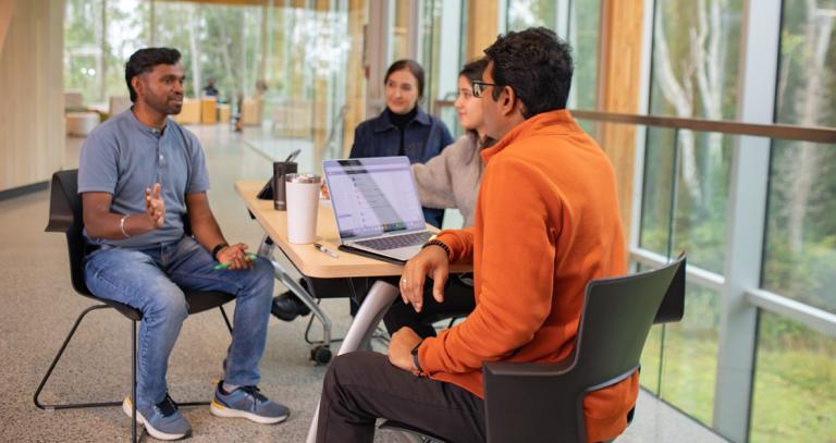 three students sit in the Sherman Jen Building chatting by the bright tree-filled windows.