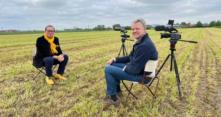 Field with two people sitting and smiling at camera with video equipment beside them.