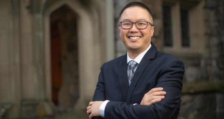 Asian man wearing glasses and a suit smiling at the camera.