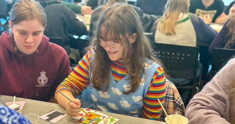 Girl painting a picture on a table, a second girl sitting next to her.